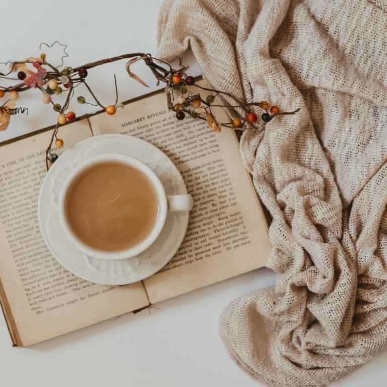 A cup of tea on an open book, surrounded by a soft, beige blanket and decorative vines.